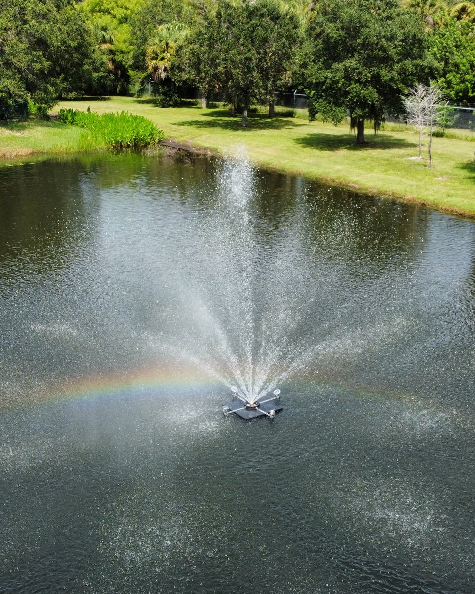 Aerial view of a floating fountain on a commercial retention pond, its spray casting a rainbow across the water