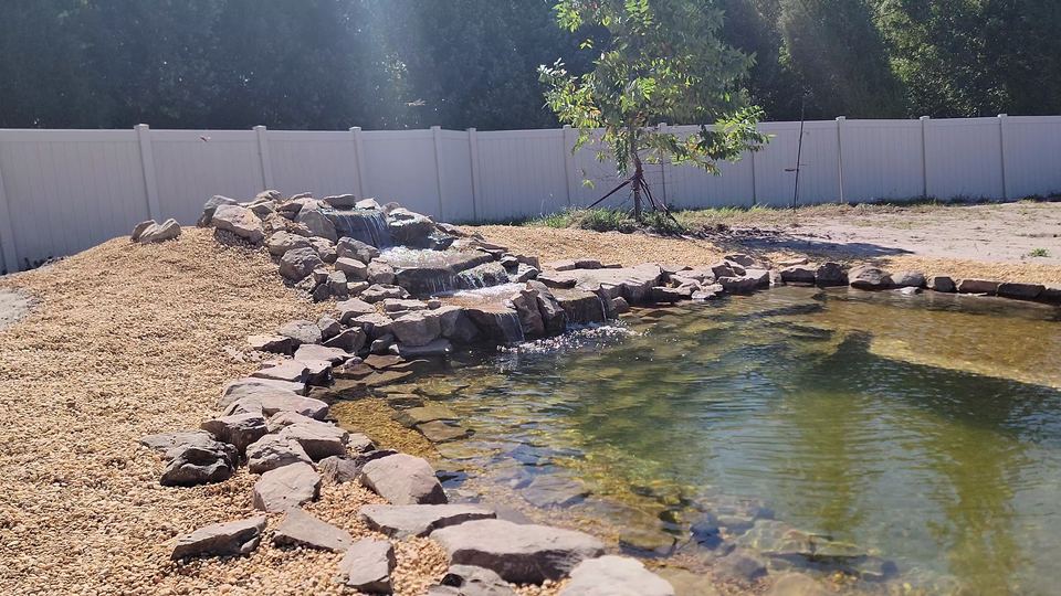 Multi-tier stone waterfall into a clear rock-edged pond with mulch bed and white fence behind