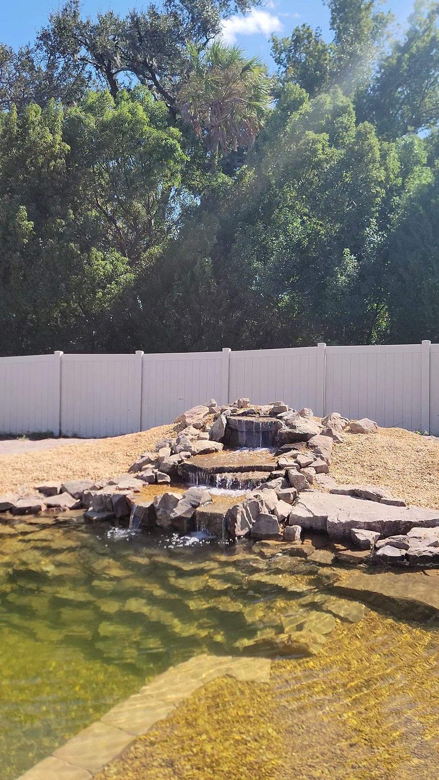 Vertical multi-tier stone waterfall pouring into a clear pond with white privacy fence and trees behind