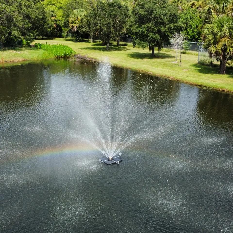 Drone view of a floating fountain spraying in a pond ringed by grass and trees