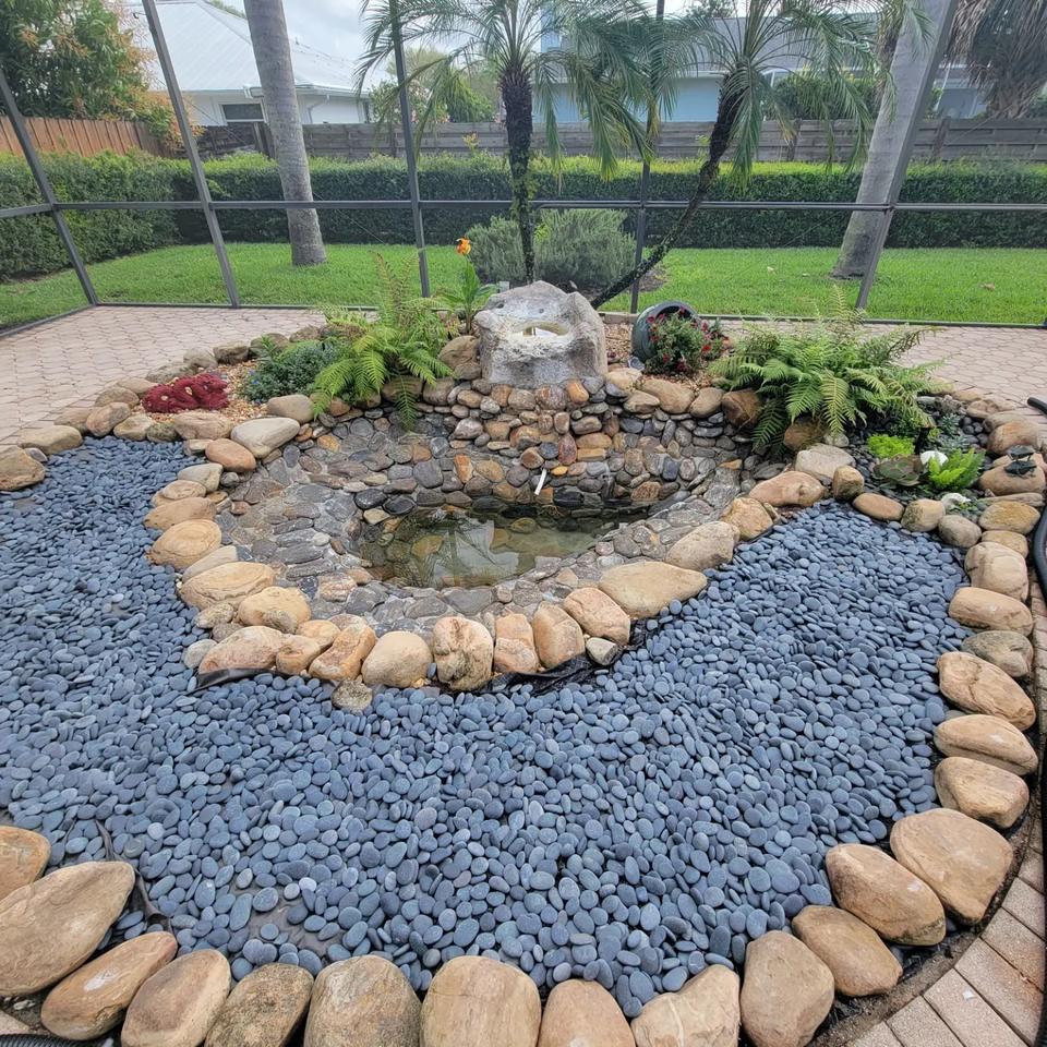 Natural stone waterfall with several spillways, edged in tan stone and set in a curving bed of black river rock with plants