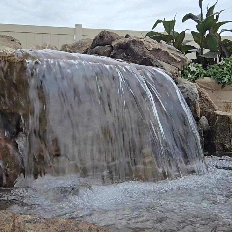 Wide sheet of water pouring smoothly off a boulder ledge into a foaming basin, with tropical plants and boulders around it