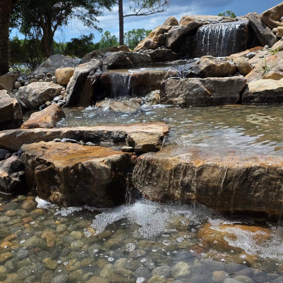 Natural stone stream cascading over flat stone ledges into a clear, shallow pool of river rock