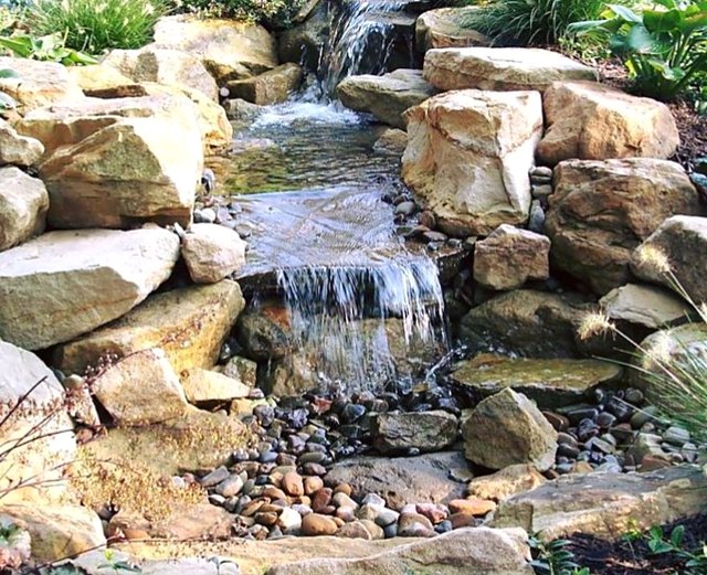 Three-tier pondless waterfall stepping down through sandstone boulders onto a bed of river rock, framed by hostas and ornamental grasses