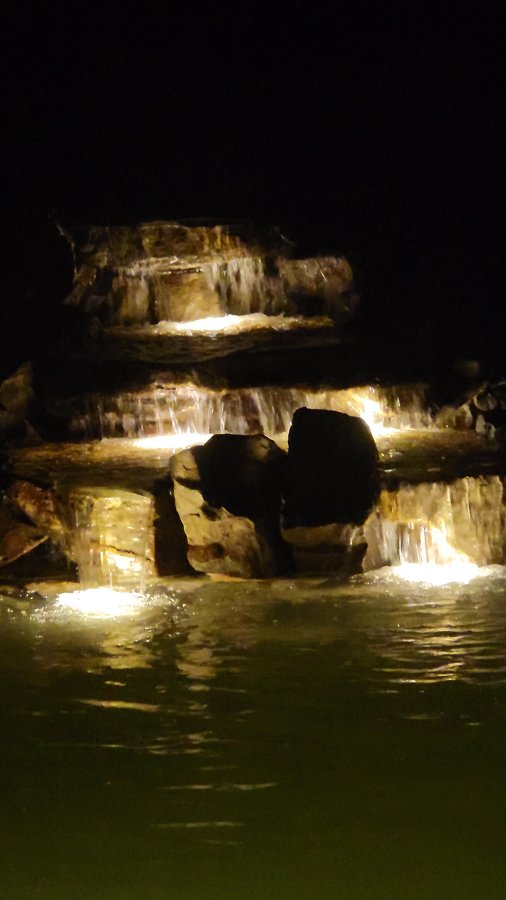 Lit multi-tier rock waterfall cascading into the swim pond at night