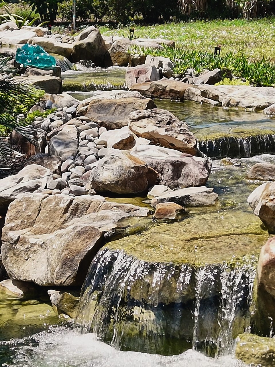 Two-tier stone waterfall spilling between boulders and river rock into a clear pond, with a green glass accent rock and lawn behind