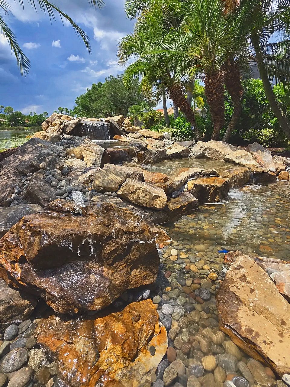 Multi-tier rock waterfall and bubbling boulder flowing into a clear pebble-bottom pond under palms, with a lake behind
